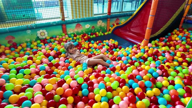 Happy girl having fun in dry paddling pool in playing centre. Little girl playing with colorful balls in playground ball pool