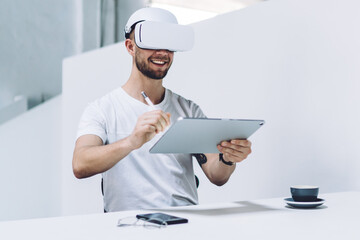 Smiling Caucasian male extends hand with digital pencil for interactive touch on tablet while sitting in interior, successful man work with 3D Model in VR headset via innovative technology application