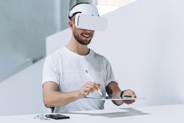 Happy man in electronic goggles sitting at office desktop and using modern digital tablet for developing imaginary environment via modern device and stylus, successful person with high technology