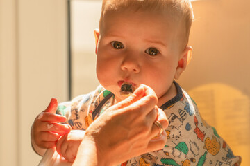 Cheerful baby boy being fed with porridge while sitting in white chair. He smiling and eating. Baby...