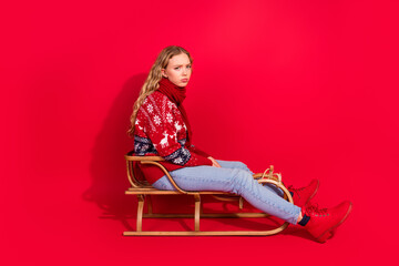 Full size photo of pretty young girl sad face sit sled isolated on christmas vibrant red color...