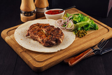 Grilled pork steak lying on tortilla on wooden board with vegetables and sauce