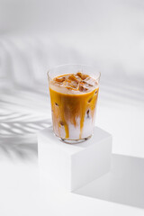 Iced caramel latte standing on white cube with palm leaf shadow