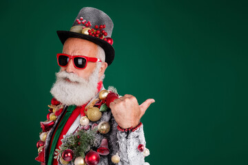 Photo of serious old cool man point empty space dressed decor balls baubles on jacket sunglass hat isolated on green color background