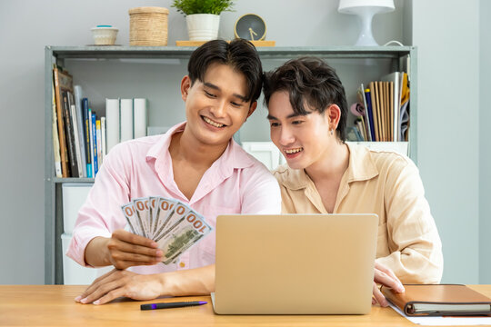 Happy two Asian men gay couple managing finances with cash banknote and laptop in home office