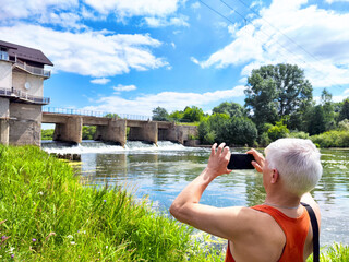 A person in a sleeveless shirt takes a picture of a river flowing over a dam on a sunny day,...