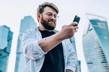 Positive young man watching funny video on mobile phone using 4G connection, smiling hipster guy in optical spectacles reading good news from networks standing on city landscape background