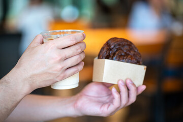 Selective focus hand of person holding appetizing baked sweet cinnamon roll in paper box and glass of ice coffee at cafe in the morning.