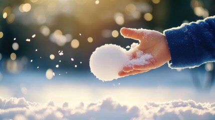 A child's hand holds a snowball, surrounded by softly falling snowflakes in a bright, winter landscape.