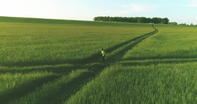 Aerial shoot of a sporty child runs through a wheat field. Evening sport training exercises at rural meadow. A happy childhood is a healthy way of life. Outdoor runing traning. Radial movement, sun