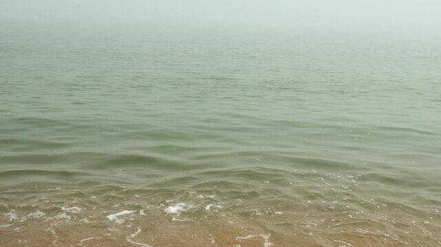 Mother-of-pearl sea surf. A quiet foggy sea, sandy-shell beach, the sun breaks through the fog and colors the crest of the rolling waves
