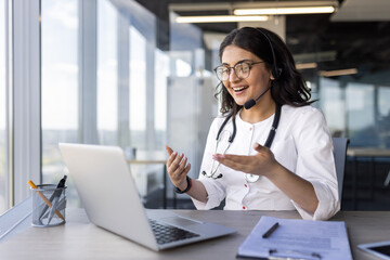 A young female doctor with a headset talking to a patient, a clinic worker consults remotely, online support, an Indian woman uses a laptop, happily smiles and discusses the client's illness