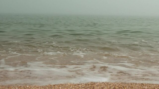 Mother-of-pearl sea surf. A quiet foggy sea, sandy-shell beach, the sun breaks through the fog and colors the crest of the rolling waves