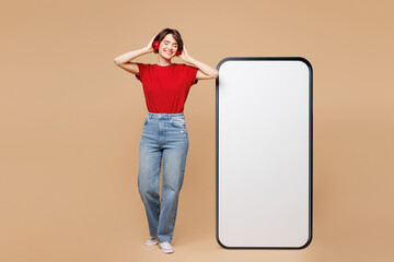 Full body young woman wear red t-shirt casual clothes big huge blank screen mobile cell phone smartphone with area listen to music in headphones isolated on plain beige background. Lifestyle concept.