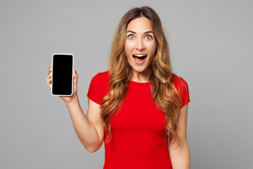 Naklejka premium Young shocked surprised happy woman she wear red t-shirt casual clothes hold in hand use mobile cell phone with blank screen workspace area isolated on plain grey background studio. Lifestyle concept.