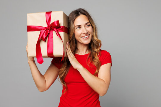Young smiling happy woman she wearing red t-shirt casual clothes hold in hand present box with gift ribbon bow look aside on area isolated on plain grey background studio portrait. Lifestyle concept.