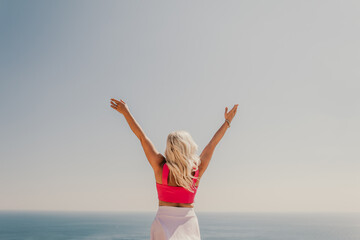 A woman in a pink top and white skirt is standing on a cliff overlooking the ocean. She is smiling and she is happy.