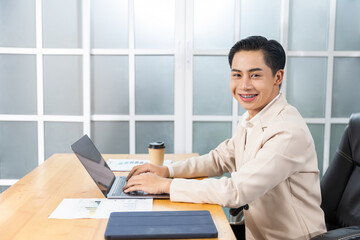 Asian businessman working in his office reviewing work, accessing planning strategy, creative thinking and innovation ideas, happy smiling sitting at desk on laptop computer professional worker.