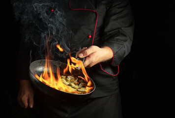 Professional chef cooking food on a frying pan with fire flames. Restaurant kitchen idea on a black...