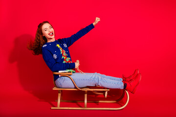 Fototapeta premium Full body photo of attractive young woman riding sledge raise fist dressed stylish blue christmas clothes theme party on red background