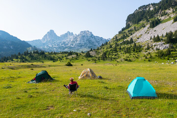 Hiker sitting on a camping chair surrounded by tents on a mountain in a beautiful valley with a view of rocky peaks in the background	