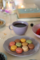 Plate of pastel macarons, cookies and chocolate, cup of tea of coffee, glass of bubble water, various berries, books and accessories on the table. Selective focus, pastel colors.