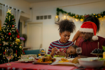 A joyful family gathers around the dinner table celebrating Christmas. The father, dressed in a Santa hat, gives a gift to the child, while everyone smiles with warmth and festive decorations.