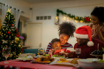A joyful family gathers around the dinner table celebrating Christmas. The father, dressed in a Santa hat, gives a gift to the child, while everyone smiles with warmth and festive decorations.