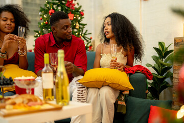 A group of friends celebrates Christmas together at home, enjoying drinks and laughter around a cozy living room. A decorated Christmas tree lights up the background, adding warmth to the holiday.