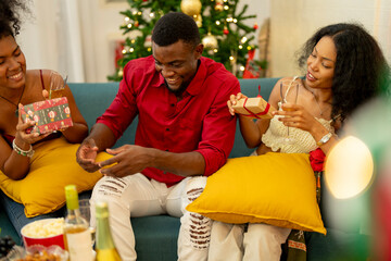 A group of friends celebrates Christmas together at home, enjoying drinks and laughter around a cozy living room. A decorated Christmas tree lights up the background, adding warmth to the holiday.
