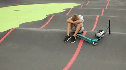 Young boy feeling sad and lonely sitting on skateboard track, hugging his knees. He has his scooter and helmet by his side