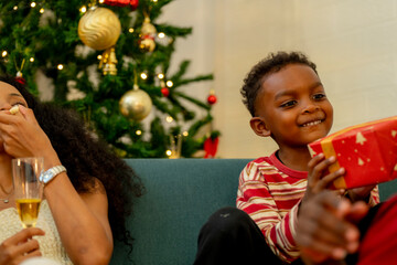 A cheerful family gathers around the Christmas tree, sharing a moment of joy as a man presents a red gift box to a child. while family members relax on the sofa, enjoying the warmth of the season.