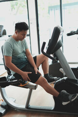 Fototapeta premium This is a kind of sport injury concept; Asian male athlete sitting on spin bicycle fitness workout and lower leg cramp and backache, Sport man massaging sore muscles during training in fitness gym.
