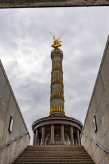 A tall, gold-covered tower with a statue of a woman on top. The sky is cloudy and the tower is...