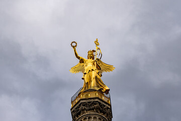Fototapeta premium A statue of a woman holding a wreath and a bird. The statue is gold and is on top of a building. The sky is cloudy and the overall mood of the image is serene and peaceful