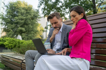 Team of diverse partners sitting at bench mature Latin business man and European business woman discussing project on laptop at park. Two colleagues of professional business people working together.