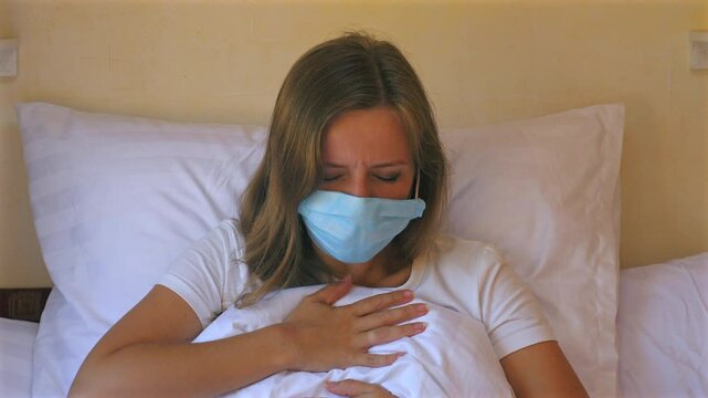 Woman wearing face mask lying in bed and holding her chest, hard coughing, depicting illness and discomfort, Health and Wellness.