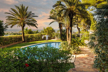 A man of retirement age in a white robe by the pool against an exotic landscape with palm trees and a pool enjoying the setting sun.
