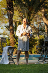 A man of retirement age in a white robe by the pool against an exotic landscape with palm trees and a pool enjoying the setting sun.
