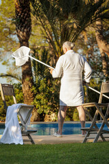 A man of retirement age in a white robe by the pool against an exotic landscape with palm trees and a pool enjoying the setting sun.
