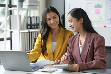 Two young businesswomen collaborate in a vibrant office, working on a laptop with bright smiles, discussing data