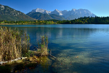 Schilf steht am Ufer eines Türkisblauen Bergsee und der Blick reicht zu einen herbstlichen...