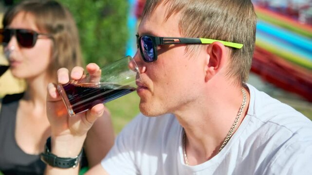 Couple enjoying soda at street restaurant sitting in sunglasses against backdrop of amusement rides and carousels spending weekend in city park.