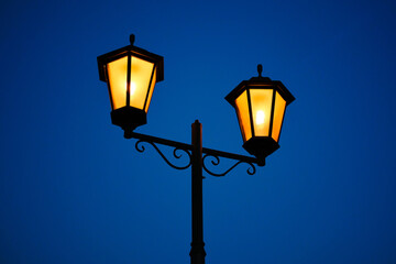 Retro style street lantern with bright yellow light against the evening deep blue sky. Vintage outdoors lamp.