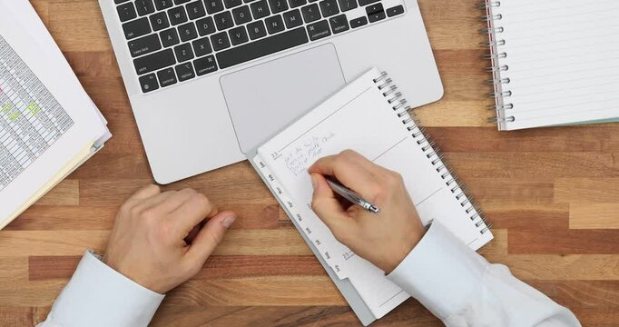 Businessman writing in notepad at wooden table with laptop and papers top view