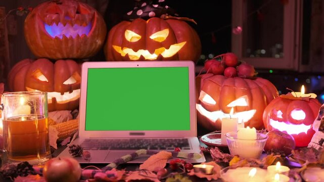 Halloween holiday background, laptop with green screen, place for ad offers, with may carved pumpkins Jack O Lanterns and flashing light, candles, colorful garlands.