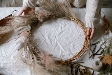 Hands holding dried grass, herbs, leaves, ribbon and scissors on rustic table. Woman making stylish autumn wreath. Fall decor and arrangement in home.