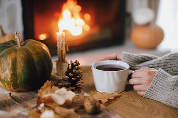 Autumn hygge. Hands holding cup of tea and pumpkin, autumn leaves on table on background of warm fireplace. Cozy fall in living room. Thanksgiving and Halloween.