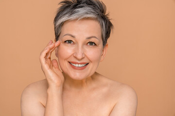 Beauty portrait of an attractive sensual mature topless woman with short gray hair standing isolated over beige background posing looking at camera. Antiage skincare cosmetology concept