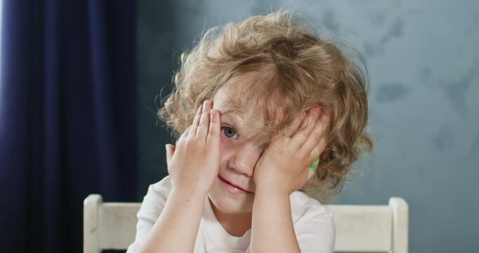 A blond child with curls closes his eyes with his palms, a portrait of a shy and timid child.